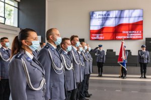 policjanci ustawienie w szeregu na tle pocztu sztandarowego i wyświetlonej na telebimie flagi Polski
