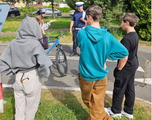 Policjant rozmawia z rowerzystką, z boku stoją3 uczniowie, którzy słuchają ich rozmowy