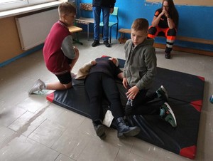 Uczniowie udzielają pierwszej pomocy symulantowi, który leży na materacu- ratownik medyczny kontroluje ich działania