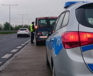 policjant dokonuje kontroli pojuazdu na drodze. Na zdjęciu widać tylny zderzak i lampę radiowozu, a w tle jezdnię.