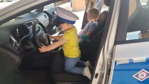 chłopczyk w białej czapce policyjnej siedzi na siedzeniu kierowcy w radiowozie, obok na siedzeniu pasażera inny chłopczyk siedzi