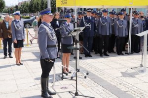 Policjantka stoi przed mównicą i czyta rozkazy personalne. Obok dowódca uroczystości, za nią zaproszeni goście