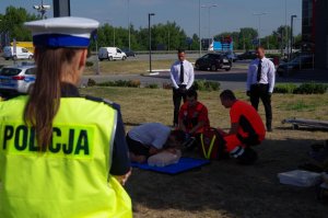 policjantka stoi tyłem do fotografującego w tle pracownicy pogotowia ratunkowego i zakładu pogrzebowego