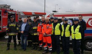 grupa osób - policjanci strażacy, ratownicy medyczni pracownicy zakładu pogrzebowego na tle karetki pogotowia i radiowozu straży pożarnej