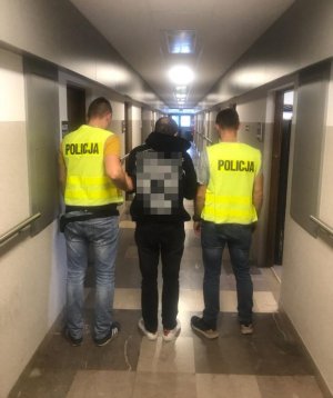 policjanci w kamizelkach odblaskowych stoją na korytarzu z zatrzymanym mężczyzną