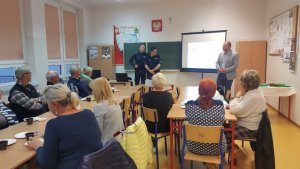 grupa ludzi, którzy siedzą przy stole w sali, przy tablicy dwóch policjantów