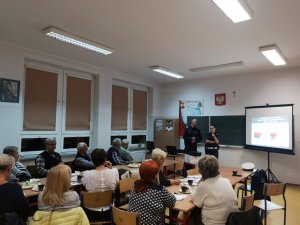 policjant z policjantką, którzy stoją przy tablicy, w sali grupa osób siedzących przy stołach