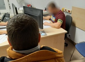 mężczyzna siedzi tyłem do fotografującego na krześle, w tle przy biurku siedzi policjant w ubraniu cywilnym