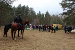 polana leśna, grupa ludzi przy ognisku w oddali dwa konie policyjne