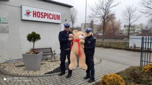 naczelnik Ruchu Drogowego wraz z policjantką trzymają dużego miśka - w tle budynek Hospicjum