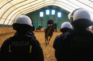 policjanci z SPPP stoją tyłem do fotografa, między nimi w tle jeźdźcy na koniach