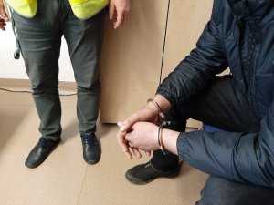 mężczyzna z kajdankami na rękach siedzi na krześle, przed nim stoi policjant w kamizelce odblaskowej