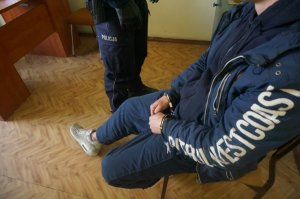 policjant stoi przy siedzącym na krześle zatrzymanym