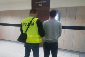 policjant w kamizelce odblaskowej chwytem transportowym trzyma podejrzanego o usiłowanie zabójstwa przed salą sądową