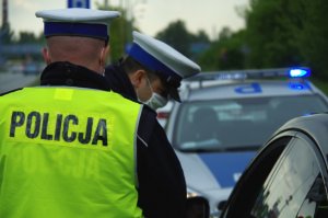 policjanci z ruchu drogowego w białych czapkach i odblaskowych kamizelkach kontrolują kierującego pojazdem osobowym