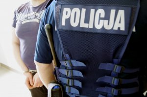 policjant umundurowany a w tle zatrzymany z kajdankami na rękach trzymanych z przodu