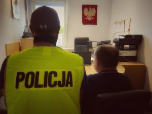 mężczyzna w kamizelce z napisem Policja oraz drugi meżczzyna skierowani tyłem do obiektywu