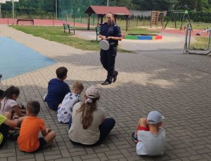 policjantka trzyma w ręku białą czapkę i opowiada o bezpieczeństwie na drodze grupie dzieci