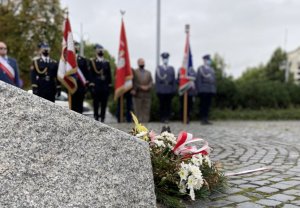 na pierwszym planie wiązanka pod pomnikiem, w tle poczet sztandarowy Komendy Miejskiej Policji w Częstochowie