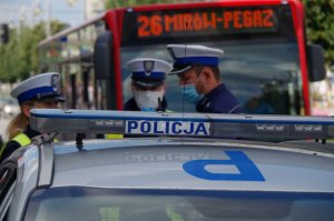 na pierwszym planie napis policja na radiowozie, w tle 3 policjantów w białych czapkach na tle autobusu rozmawia ze sobą