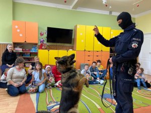 Policjant pokazującyc pkaz tresury psa. Pies stoi na dwóch łapach przed policjantem w tyle dzieci sidzace na materacu.