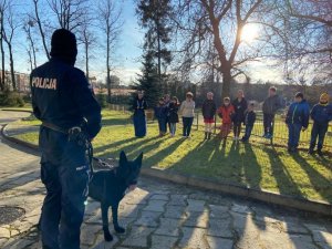 Policjant z psem na smyczy w tle dzieci stojące przed nimi.