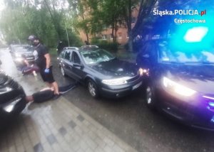 radiowóz policyjny i samochód, którym poruszali się przestępcy stoją na drodze. Nieumundurowany policjant pilnuje leżących na ziemi przestępców