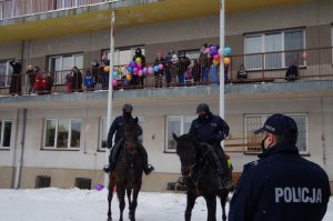 policjant stoi tyłem - widać napis Policja na jego kurtce, w tle 2 konie służbowe i dzieci na balkonie