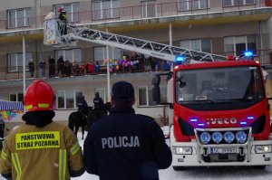 policjant i strażak stoją obok siebie i patrzą na balkon, gdzie są dzieci i biskup z drugim strażakiem w koszu na pojeździe strażackim