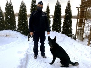 policjant stoi obok przy jego nodze siedzi czarny pies rasy owczarek niemiecki
