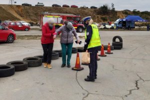 uczestniczka pikniku ma założone alkogole, pokonuje w nich tor przeszkód ułożony z pachołków. Obok trzyma ją za ramię druga kobieta a w pobliżu stoi umundurowana policjantka