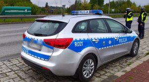 dwóch policjantów w tle radiowozu kontrolują prędkość przejeżdżających samochodów przy użyciu ręcznego miernika prędkości