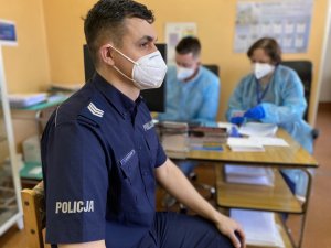 policjant oczekuje na szczepienie siedząc na krześle, w tle pielęgniarki przy biurku