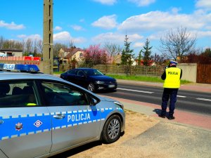 policjant na tle radiowozu trzyma w dłoni ręczny miernik prędkości, po drodze jedzie czarny samochód