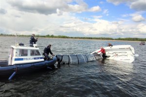 Policyjna łódka na które stoi dwóch policjantów, jeden z nich trzyma żaglówkę.