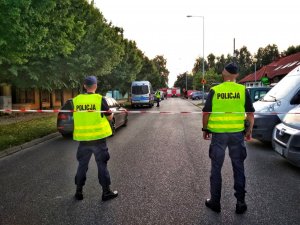 dwóch policjantów w kamizelkach odblaskowych stoi tyłem do fotografującego. W tle radiowozy i wozy strażackie