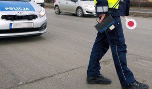 policjant trzyma w ręce urządzenie do pomiaru prędkości oraz tarcze do zatrzymywania pojazdów - w tle radiowóz