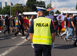 policjanci maszerują drogami miasta zabezpieczając Marsz Równości - w tle kolorowa flaga uczestników zgromadzenia