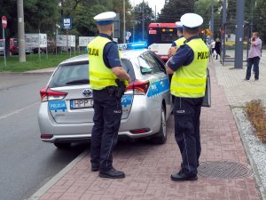 3 policjantów w kamizelkach odblaskowych stoi przy radiowozie, w tle autobus
