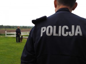 napis policja na mundurze policjanta, w tle policjant z psem służbowym podczas pokazu posłuszeństwa