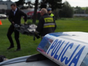 napis policja na radiowozie, w tle sprawca wykroczenia i pracownik zakładu pogrzebowego podczas przenoszenia noszy