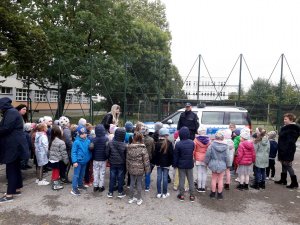 policjant stoi przed radiowozem, wokół grupa przedszkolaków na parkingu wewnętrznym placówki