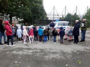 policjant stoi przed radiowozem, wokół grupa przedszkolaków na parkingu wewnętrznym placówki