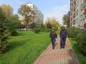 Teren placu zabaw wewnątrz osiedla blokowisk, chodnikiem idą strażniczka miejska oraz policjant