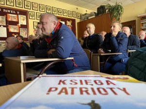 policjanci w ubraniach cywilnych oglądają występ w szkole, na pierwszym planie na ławce leży podręcznik w języku polskim