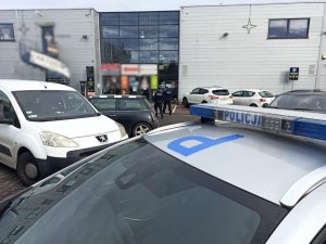 radiowóz ustawiony na parkingu przed sklepem, w tle dwóch policjantów wchodzi do sklepu