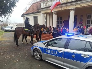 policyjny radiowóz i dwóch policjantów na koniach stoją przed wejściem do przedszkola, przy wejściu stoi grupa przedszkolaków