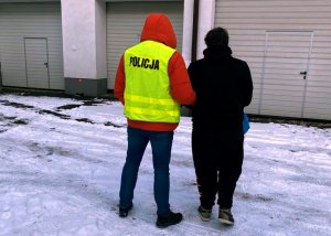 policjant w kamizelce odblaskowej trzyma pod rękę zatrzymanego - stoją tyłem do fotografującego