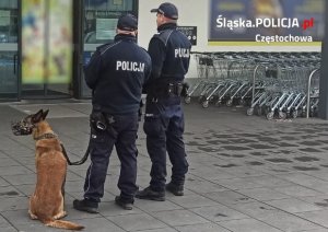 dwóch policjantów z psem słuzbowym stoi przed galerią handlową - zdjęcie poglądowe