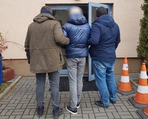 zatrzymany mężczyzna jest prowadzony do budynku przez dwóch nieumundurowanych policjantów, prowadzących go z dwóch stron pod ramię